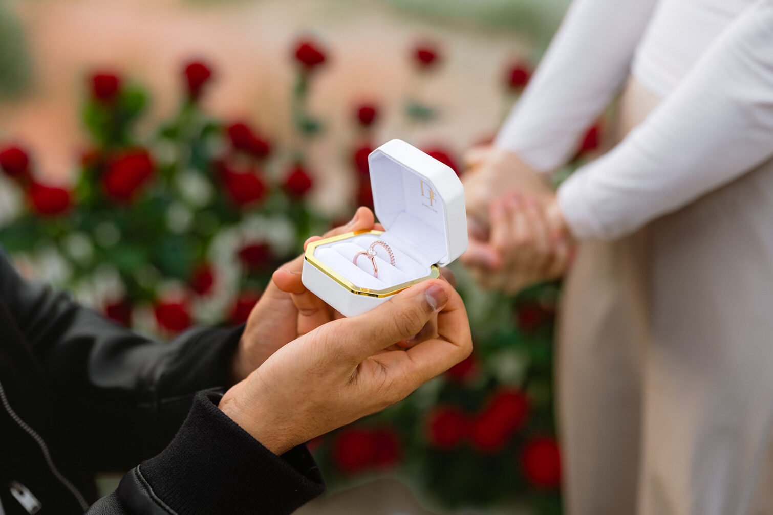 Engagement ring in an open box with red roses from the proposal setup in the background